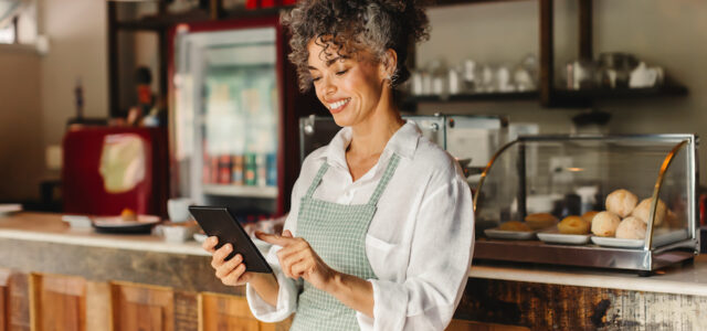 Happy business owner using a digital tablet in her cafe. Mature female cafe owner smiling cheerfully while standing in front of the counter. Successful entrepreneur running her business online.