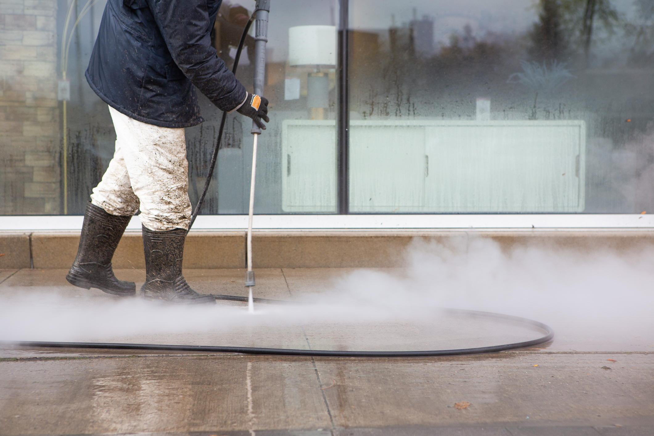 Worker power washing the sidewalk wearing black rubber boots and white pants