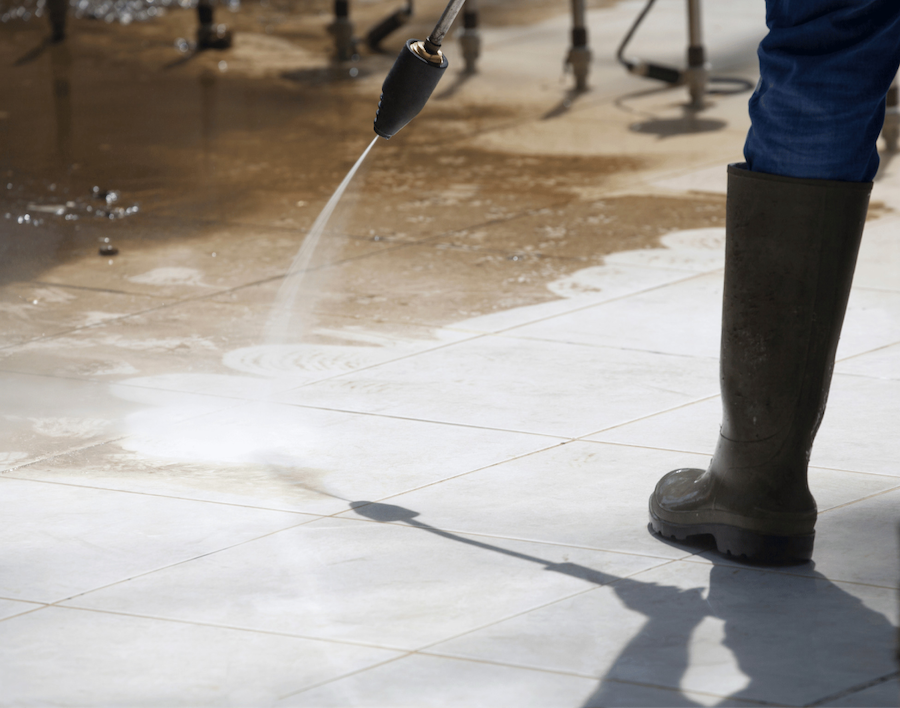 A person wearing large black rain boots pressure washes outdoor concrete tiles. 