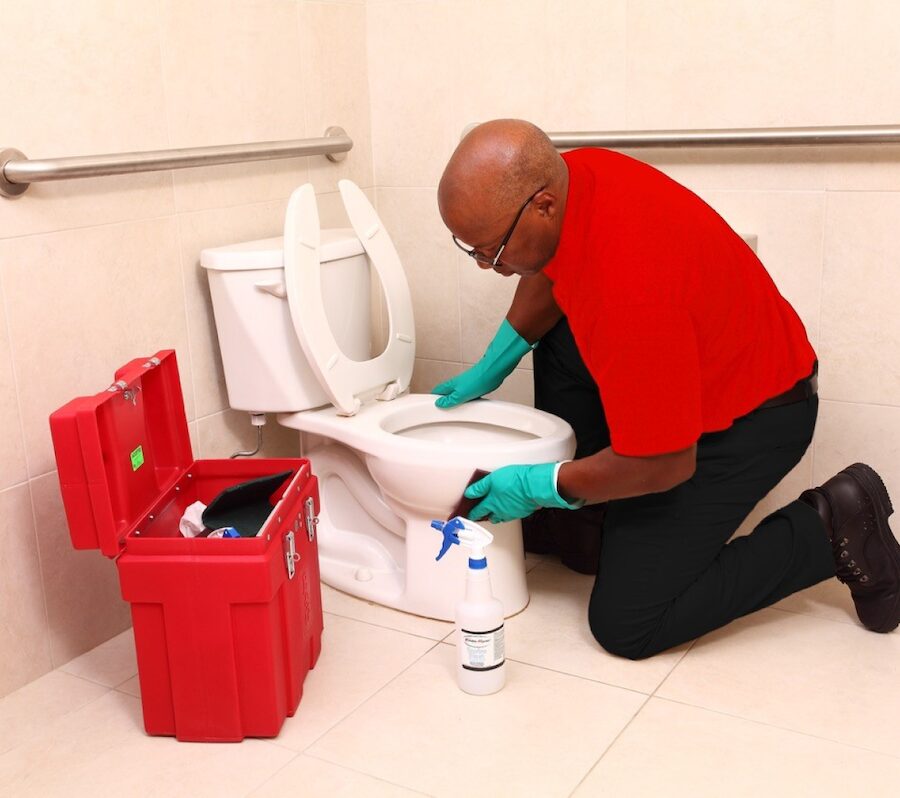 Enviromaster worker in red on hand and knees cleaning a public restroom toilet.